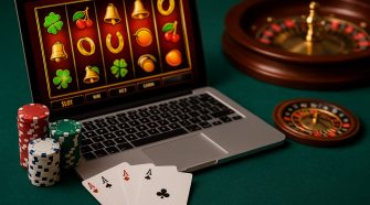 A high-resolution landscape photo showing a laptop with a slot game on screen, poker chips stacked beside it, three aces on the table, and a roulette wheel in the background.