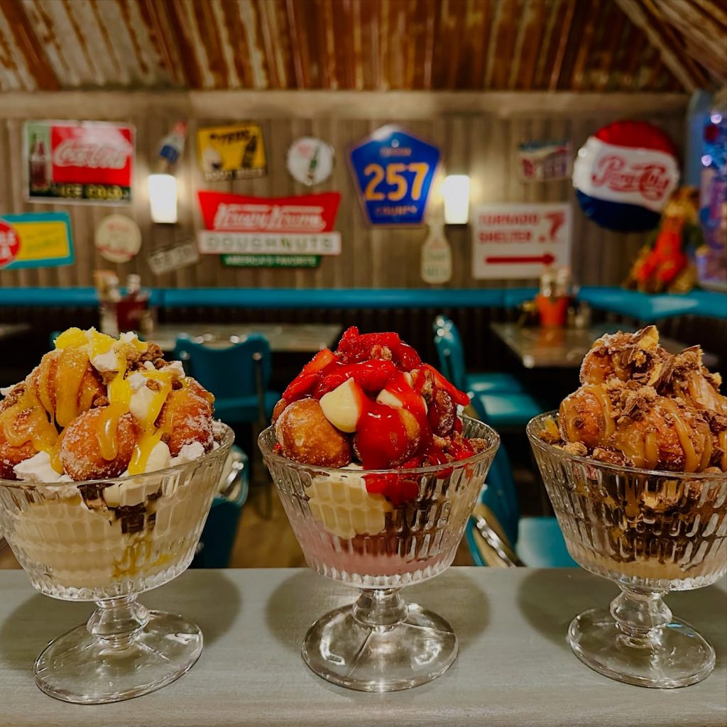 Three large glass dessert bowls filled with indulgent sundaes topped with doughnut bites, sauces, fruit, and cream, set against a retro American diner backdrop with vintage signs and teal chairs.
