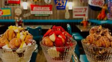 Three large glass dessert bowls filled with indulgent sundaes topped with doughnut bites, sauces, fruit, and cream, set against a retro American diner backdrop with vintage signs and teal chairs.