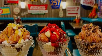 Three large glass dessert bowls filled with indulgent sundaes topped with doughnut bites, sauces, fruit, and cream, set against a retro American diner backdrop with vintage signs and teal chairs.