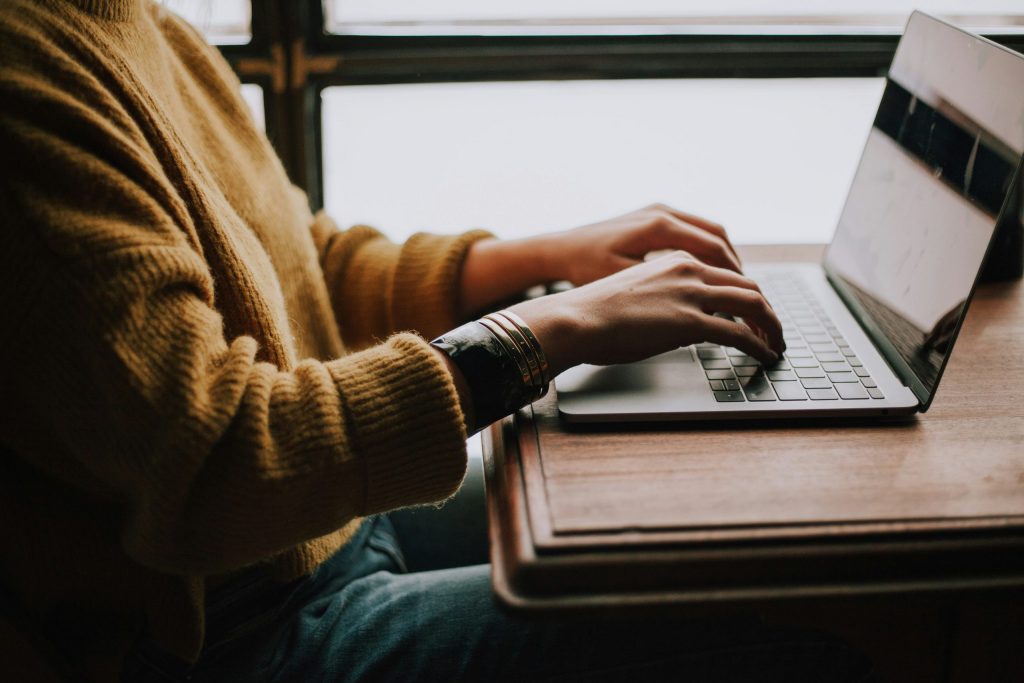 Person in a mustard yellow sweater typing on a laptop at a wooden desk near a window.
