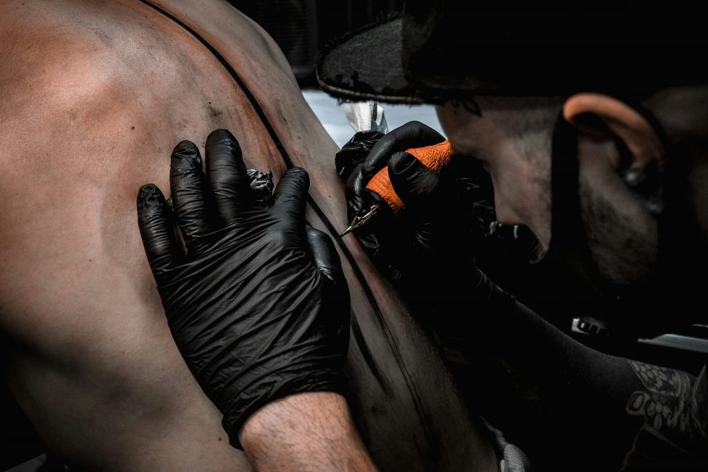 Tattoo artist wearing black gloves working on a client’s back with a tattoo machine in a studio.