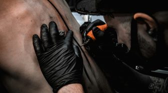 Tattoo artist wearing black gloves working on a client’s back with a tattoo machine in a studio.