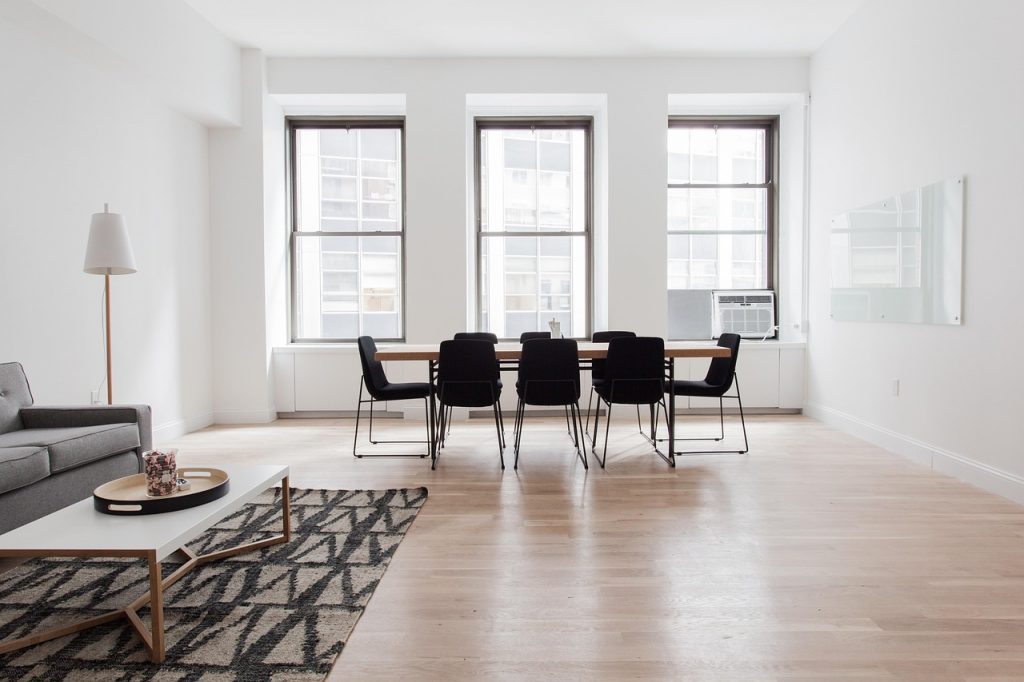A bright, minimalist living and dining space featuring light wooden floors, a simple grey sofa, a modern white coffee table with a geometric rug, and a sleek wooden dining table surrounded by black chairs in front of large windows.