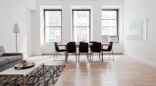 A bright, minimalist living and dining space featuring light wooden floors, a simple grey sofa, a modern white coffee table with a geometric rug, and a sleek wooden dining table surrounded by black chairs in front of large windows.