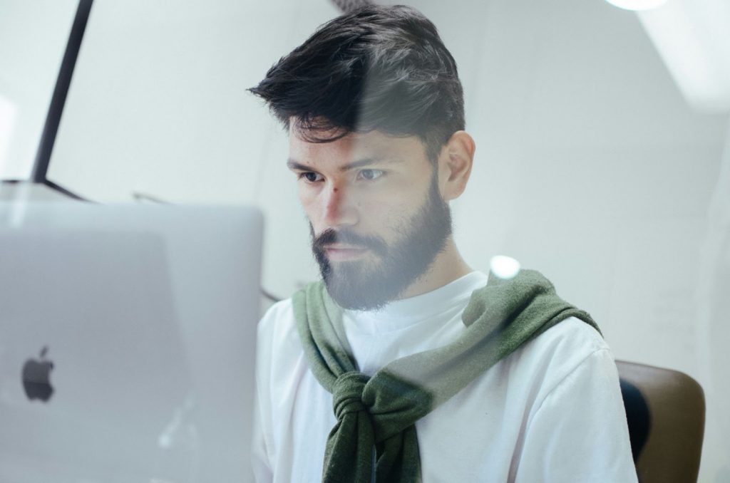 A man with a beard and dark hair is sitting at a desk, focused on his computer screen. He is wearing a white shirt with a green jumper draped over his shoulders, working in a bright, modern setting.