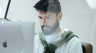 A man with a beard and dark hair is sitting at a desk, focused on his computer screen. He is wearing a white shirt with a green jumper draped over his shoulders, working in a bright, modern setting.