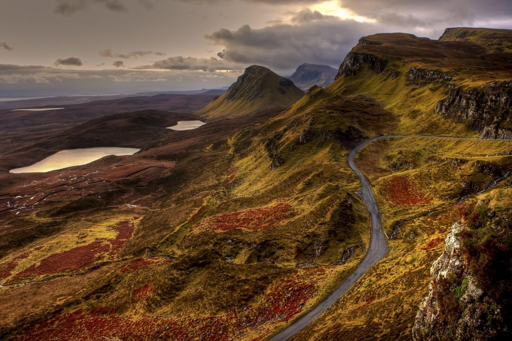 A winding mountain road cutting through the dramatic, rugged landscape of the Quiraing on the Isle of Skye, with moody skies and rich autumnal colours across the hills.
