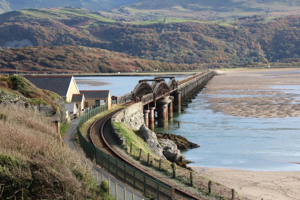 A scenic railway track curving along the coast toward a historic wooden bridge crossing the water, surrounded by mountains and cottages in rural Wales.