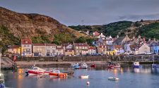 Picturesque view of Staithes harbour in North Yorkshire at dusk, with colourful fishing boats moored in calm water and traditional stone cottages nestled against the hillside.