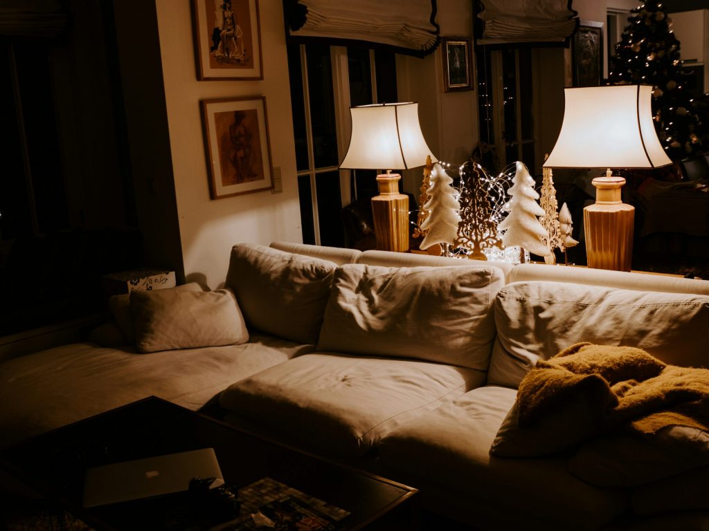 A warm and inviting living room softly lit by two table lamps, featuring a cream-coloured sofa with cosy cushions and a mustard throw. Decorative wooden Christmas trees and soft fairy lights add a festive glow, creating a relaxed, homely atmosphere.