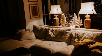 A warm and inviting living room softly lit by two table lamps, featuring a cream-coloured sofa with cosy cushions and a mustard throw. Decorative wooden Christmas trees and soft fairy lights add a festive glow, creating a relaxed, homely atmosphere.