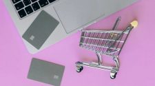 Small metal shopping trolley next to a laptop and two bank cards on a pink background, representing online payments and digital shopping.
