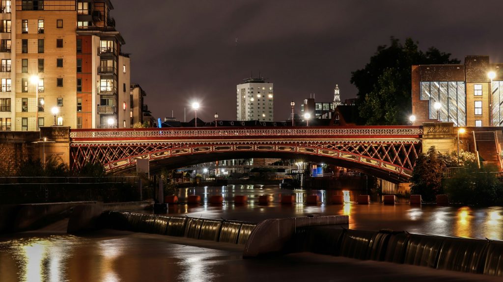 Night-time view of Leeds city centre in Yorkshire, with an illuminated historic bridge over the river and modern apartments and office buildings in the background.