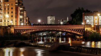 Night-time view of Leeds city centre in Yorkshire, with an illuminated historic bridge over the river and modern apartments and office buildings in the background.