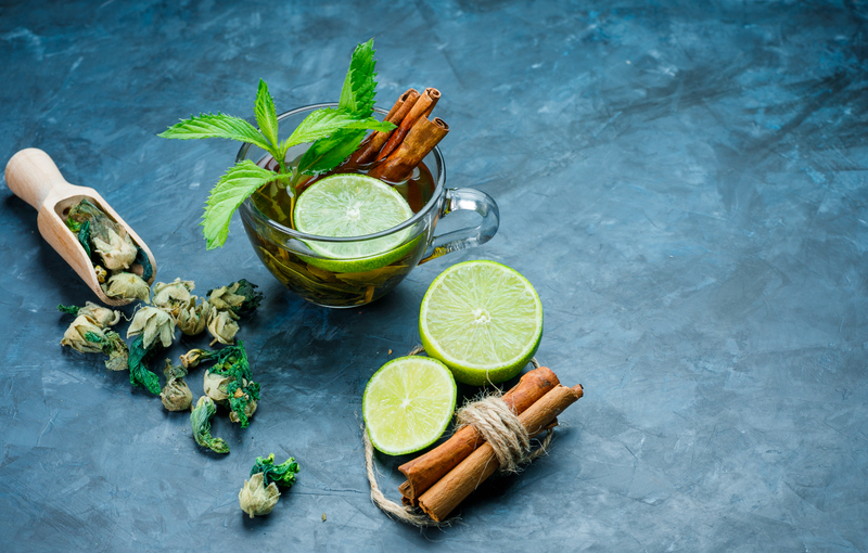Glass cup of herbal tea garnished with lime slices, mint leaves and cinnamon sticks on a blue stone background, with dried herbs and fresh lime beside it.