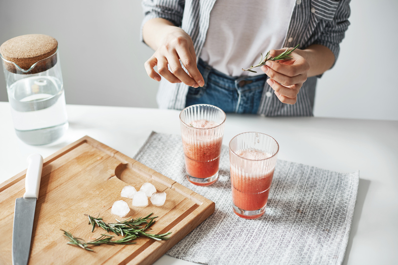 Person preparing two tall glasses of pink iced drink, adding fresh rosemary sprigs beside a wooden chopping board with ice cubes and herbs in a bright modern kitchen.