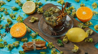 Clear cup of herbal tea on a wooden board surrounded by dried flowers, cinnamon sticks, star anise and halved citrus fruits on a bright turquoise surface.