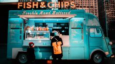 Woman and child ordering from a turquoise fish and chips food truck in an urban street setting at dusk.