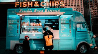 Woman and child ordering from a turquoise fish and chips food truck in an urban street setting at dusk.