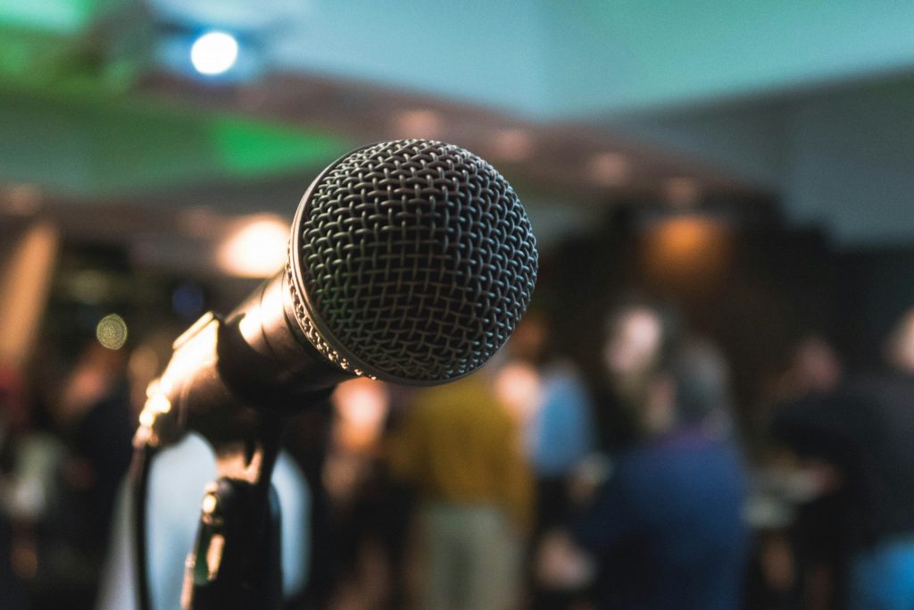 Close-up of a microphone on stage at an open mic night, with a blurred crowd of people in the background.