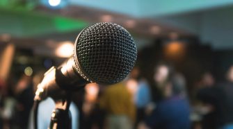 Close-up of a microphone on stage at an open mic night, with a blurred crowd of people in the background.