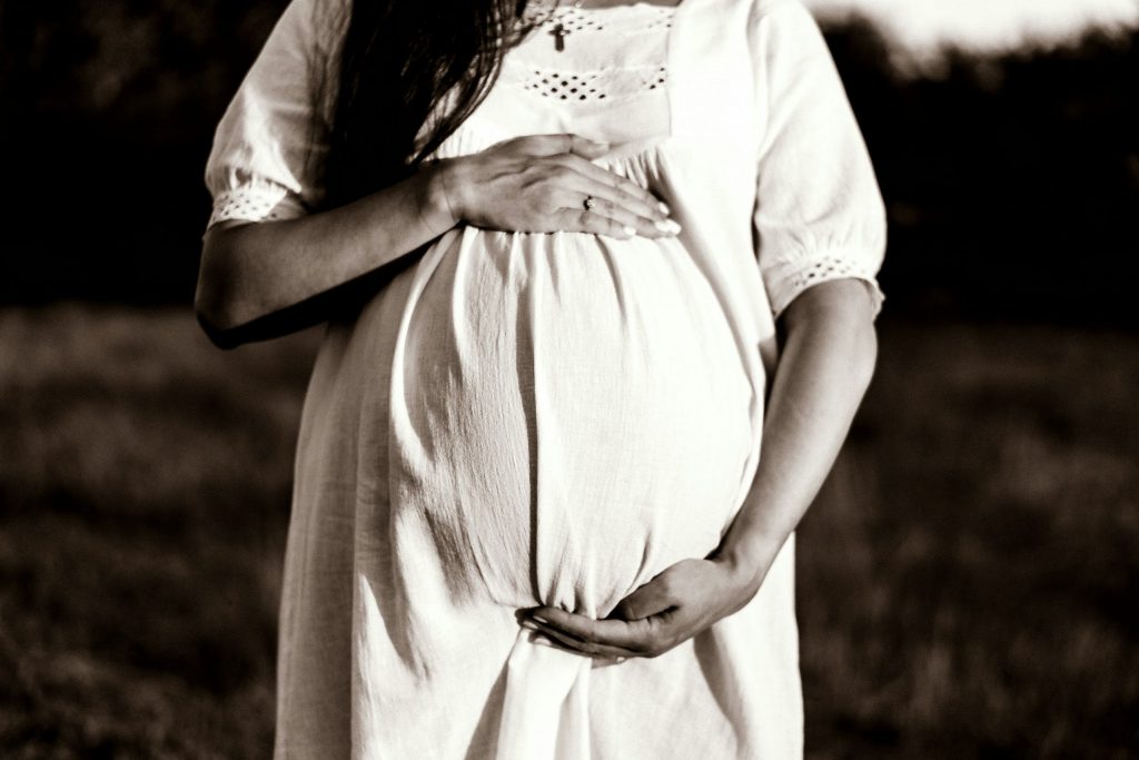 Close-up of a pregnant woman in a white dress standing outdoors, hands gently cradling her baby bump.