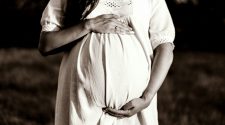 Close-up of a pregnant woman in a white dress standing outdoors, hands gently cradling her baby bump.