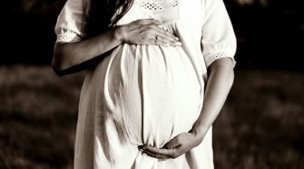 Close-up of a pregnant woman in a white dress standing outdoors, hands gently cradling her baby bump.