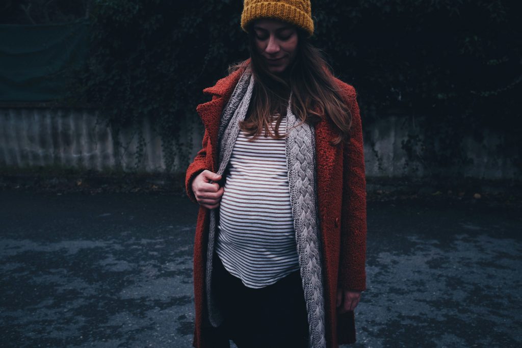 Pregnant woman in a striped top, orange coat and yellow beanie looking down at her baby bump while standing on a quiet street.