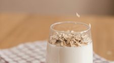 Hand sprinkles rolled oats into a glass of oat milk on a red-and-white check cloth, illustrating simple plant-based milk.