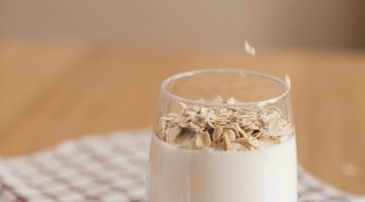 Hand sprinkles rolled oats into a glass of oat milk on a red-and-white check cloth, illustrating simple plant-based milk.