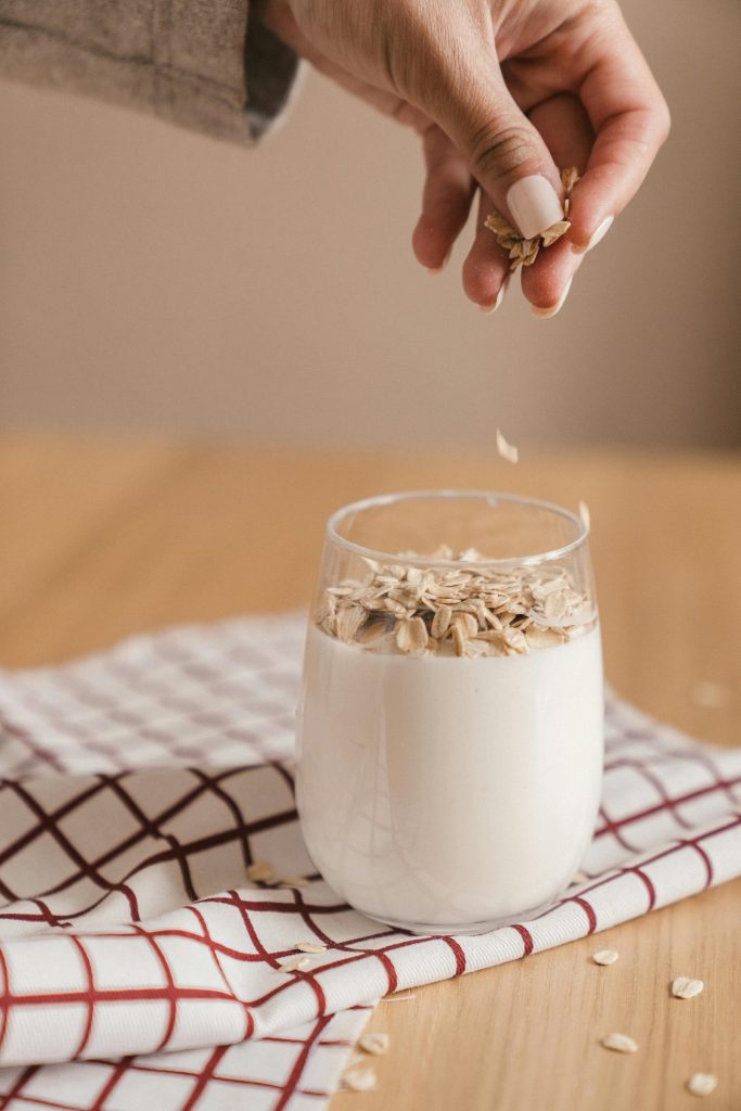 Hand sprinkles rolled oats into a glass of oat milk on a red-and-white check cloth, illustrating simple plant-based milk.