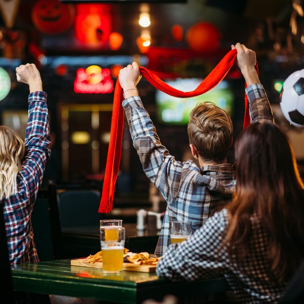 Group of football fans in a pub watching a match, with one supporter raising a red scarf while pints of beer and plates of food sit on the table in front of them.