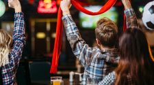 Group of football fans in a pub watching a match, with one supporter raising a red scarf while pints of beer and plates of food sit on the table in front of them.
