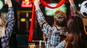 Group of football fans in a pub watching a match, with one supporter raising a red scarf while pints of beer and plates of food sit on the table in front of them.