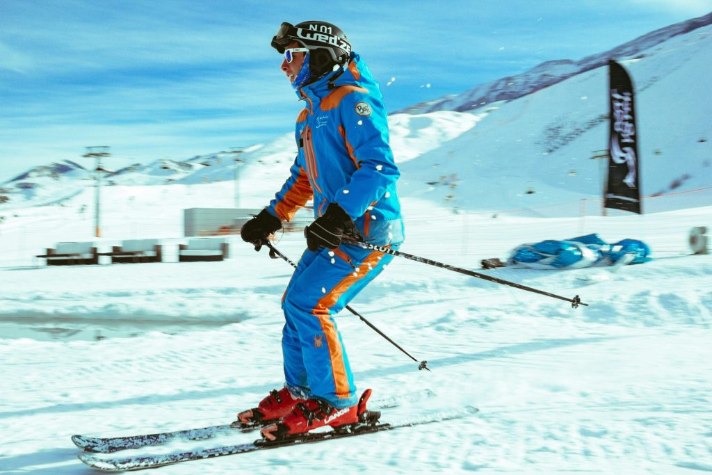 A skier in a bright blue and orange suit gliding downhill on red skis in Val Thorens, with sunny blue skies and snow-covered Three Valleys mountains in the background.