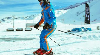 A skier in a bright blue and orange suit gliding downhill on red skis in Val Thorens, with sunny blue skies and snow-covered Three Valleys mountains in the background.