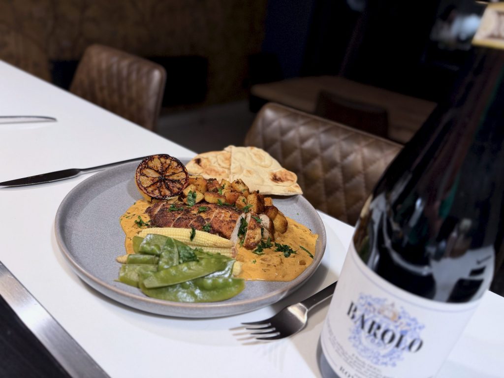 Pan-fried spiced chicken breast served on a pool of creamy Indian-style sauce with crispy diced potatoes, mange tout, baby corn and naan bread, styled on a grey plate with a bottle of red wine in a modern kitchen setting.