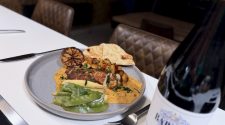 Pan-fried spiced chicken breast served on a pool of creamy Indian-style sauce with crispy diced potatoes, mange tout, baby corn and naan bread, styled on a grey plate with a bottle of red wine in a modern kitchen setting.