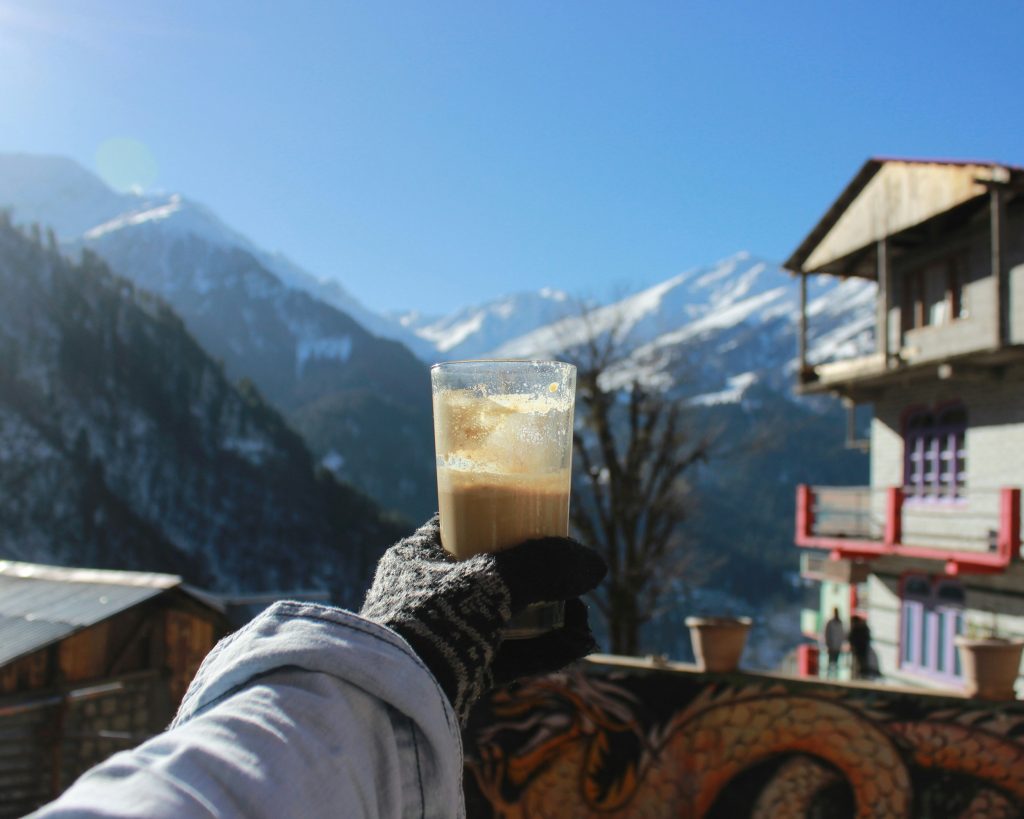 Gloved hand holding a glass of coffee on a sunny alpine terrace with snow-covered mountains and a chalet in the background.