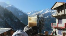 Gloved hand holding a glass of coffee on a sunny alpine terrace with snow-covered mountains and a chalet in the background.