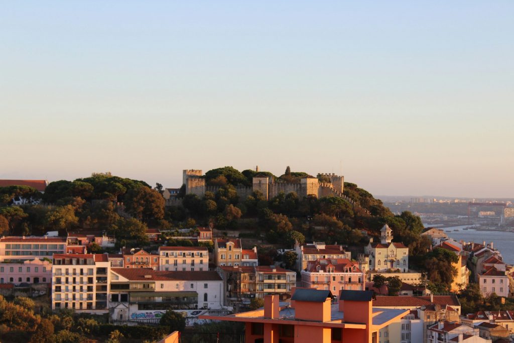 Panoramic view of Lisbon at golden hour, with São Jorge Castle on the hilltop surrounded by trees and colourful houses overlooking the river.