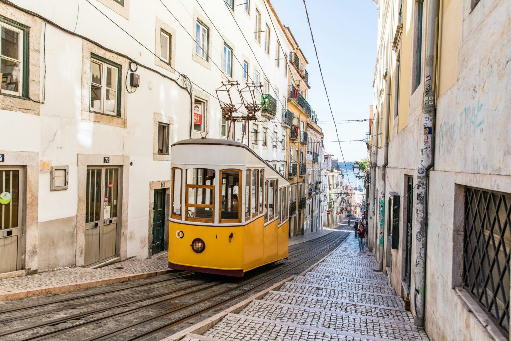 Classic yellow tram climbing a steep cobbled street in Lisbon, framed by pastel buildings and a glimpse of the Tagus River in the distance.
