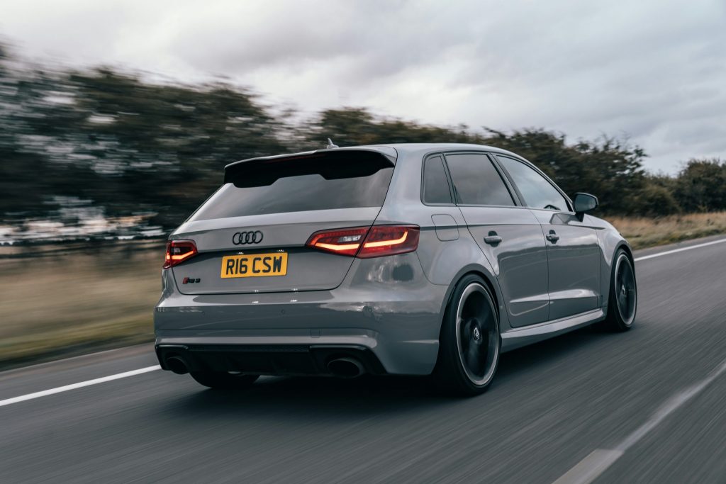Grey Audi hatchback driving on a motorway, rear three-quarter view with motion blur and overcast sky.