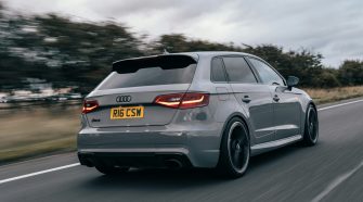 Grey Audi hatchback driving on a motorway, rear three-quarter view with motion blur and overcast sky.