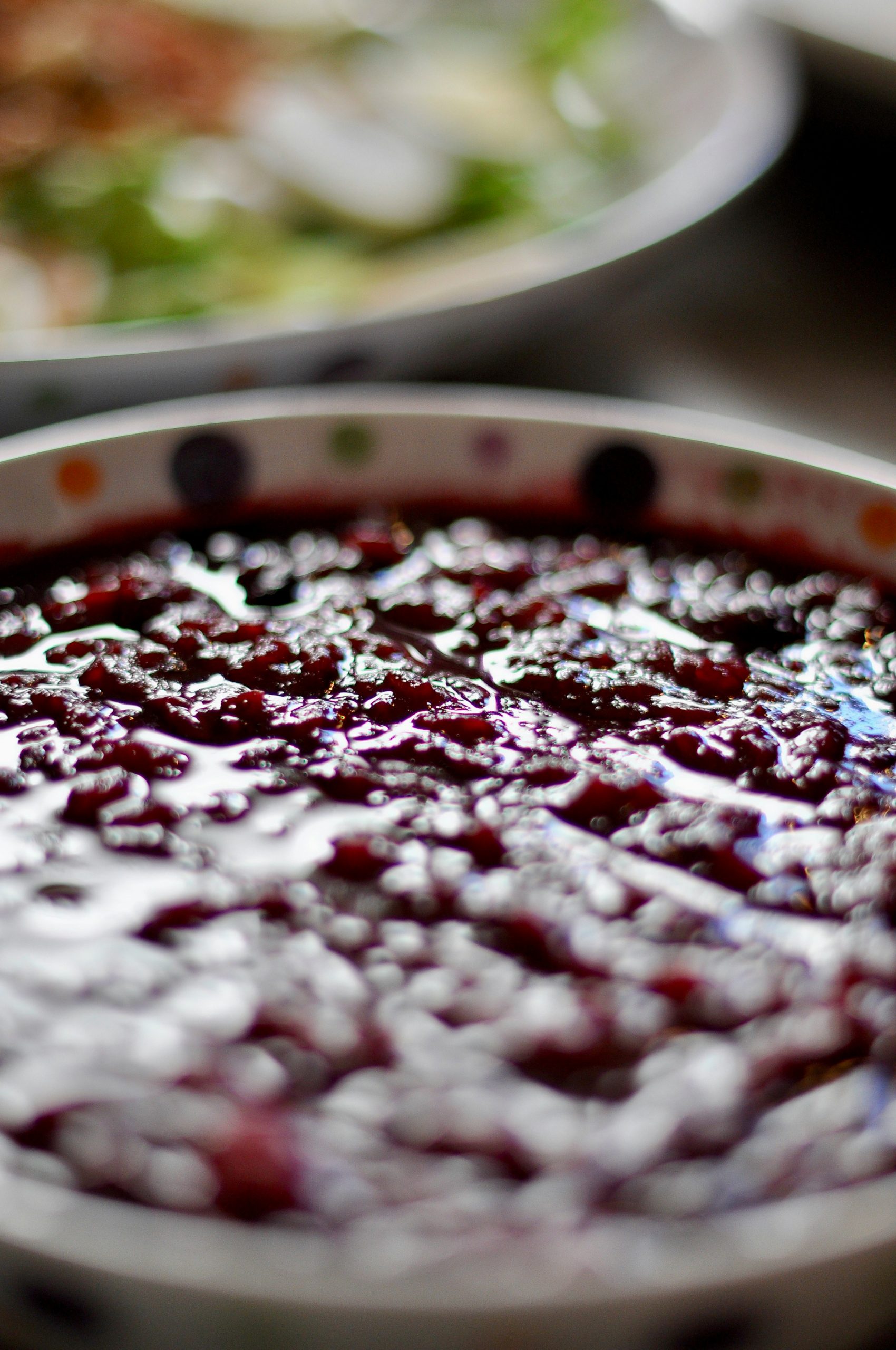 Close-up of a shallow bowl brimming with glossy, chunky cranberry sauce, with the background softly blurred to show a festive table setting.