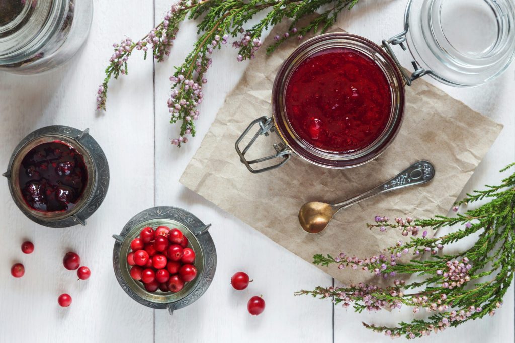 Overhead view of a glass jar filled with homemade cranberry sauce on crumpled brown paper, surrounded by fresh cranberries, berry compote and heather on a white wooden table.
