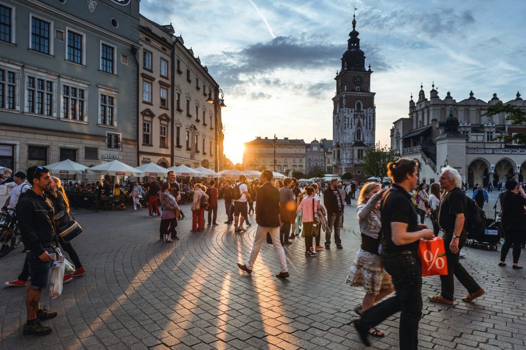 Bustling early-evening scene in Kraków’s Rynek Główny, with crowds strolling past cafés and historic townhouses as the sun sets behind the church tower.