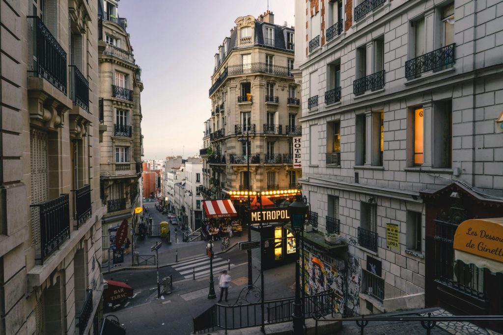 Atmospheric Parisian side street near Montmartre, lined with ornate apartment buildings, a glowing Métro sign and café awnings at dusk.